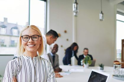 Eine Frau mit Brille lächelt in die Kamera, während im Hintergrund mehrere Menschen an einem Tisch arbeiten.