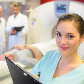 Radiologie Technikerin steht vor einem Patienten, der eine radiologische Untersuchung hat, und schaut in die Kamera.