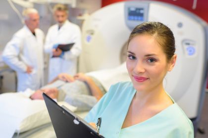 Radiologie Technikerin steht vor einem Patienten, der eine radiologische Untersuchung hat, und schaut in die Kamera.