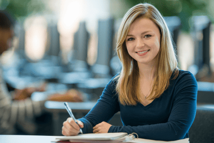 Junge Frau sitzt mit Stift und Mappe an einem Tisch und lächelt in die Kamera.