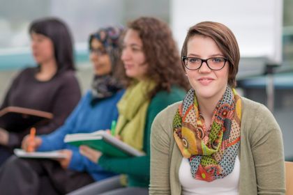 Eine junge Frau mit Brille blickt sitzend in die Kamera. Hinter ihr sitzen weitere Frauen mit Büchern in den Händen.