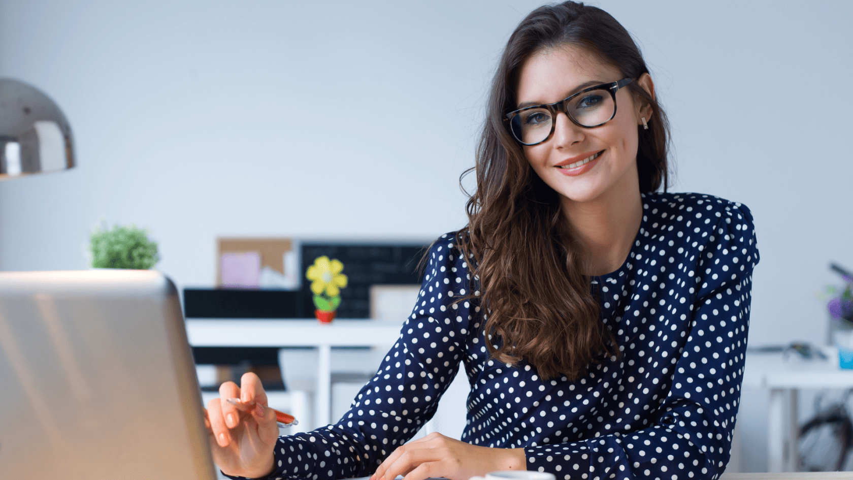 Frau mit Brille und einem Stift in der Hand sitzt hinter einem Laptop und sieht in die Kamera.