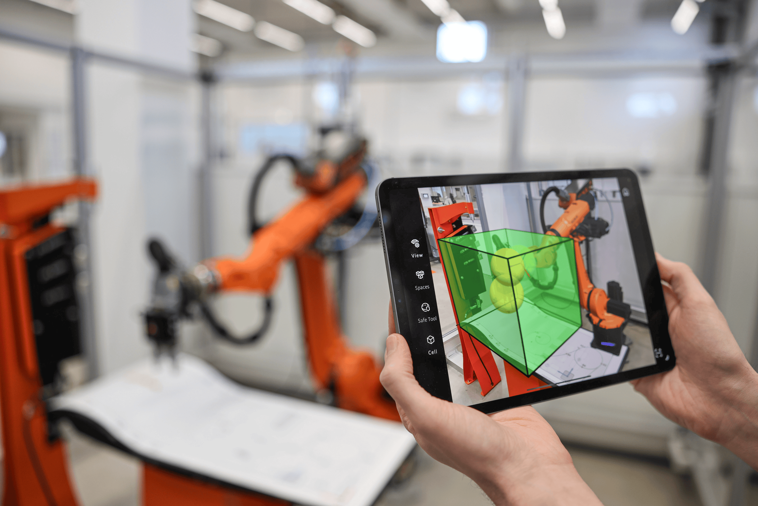 zwei Hände halten ein Tablet, das einen orangefarenen Roboter filmt