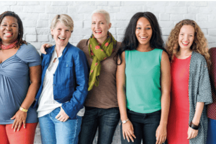 acht verschiedene Frauen stehen buntgekleidet nebeneinander vor einer Wand und lachen in die Kamera