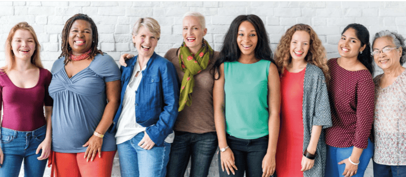 acht verschiedene Frauen stehen buntgekleidet nebeneinander vor einer Wand und lachen in die Kamera